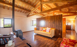 Cozy living room with wooden beams, featuring a stylish sofa, vibrant cushions, and a warm color palette, complemented by large windows that offer natural light and a view of the surroundings.