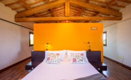 Cozy bedroom with a wooden ceiling, featuring a large bed with decorative pillows and an orange accent wall, creating a warm and inviting atmosphere. Ideal for relaxation and comfort.