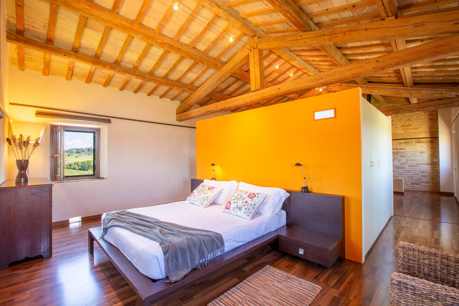 Cozy bedroom with wooden beams and an orange accent wall, featuring a comfortable bed with decorative pillows, a bedside table, and a scenic window view. Ideal for relaxation and stylish interior design. | Only on SopranoVillas