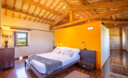 Cozy bedroom with wooden beams and an orange accent wall, featuring a comfortable bed with decorative pillows, a bedside table, and a scenic window view. Ideal for relaxation and stylish interior design.