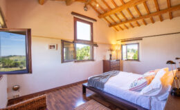 Bright and airy bedroom with wooden beams, large windows offering scenic views, a comfortable bed with decorative pillows, and a stylish wooden dresser. Ideal for a relaxing getaway.