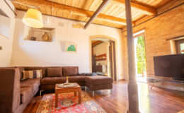 Cozy living room featuring a brown sectional sofa, wooden beams, and a flat-screen TV, with natural light streaming through the windows and a warm, inviting atmosphere.