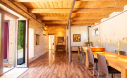 Spacious dining area with wooden beams and polished hardwood floors, featuring a long dining table and modern decor, illuminated by natural light from large windows and doors.