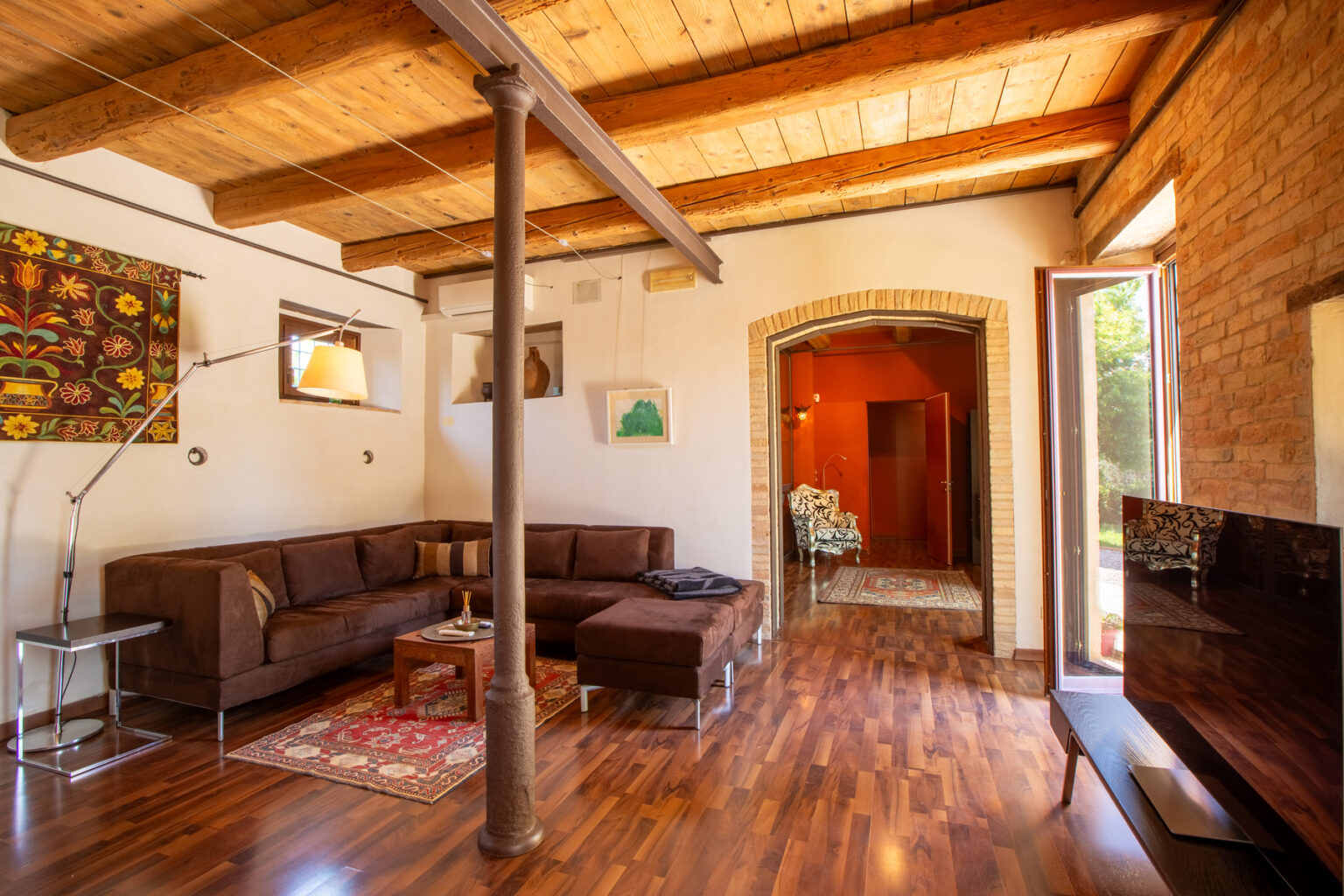 Cozy living room featuring a brown sectional sofa, wooden ceiling beams, and a stylish TV stand, with vibrant artwork on the walls and natural light streaming in from an open door. | Only on SopranoVillas