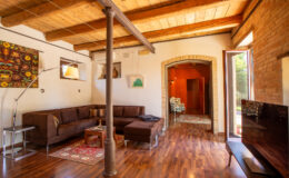 Cozy living room featuring a brown sectional sofa, wooden ceiling beams, and a stylish TV stand, with vibrant artwork on the walls and natural light streaming in from an open door.