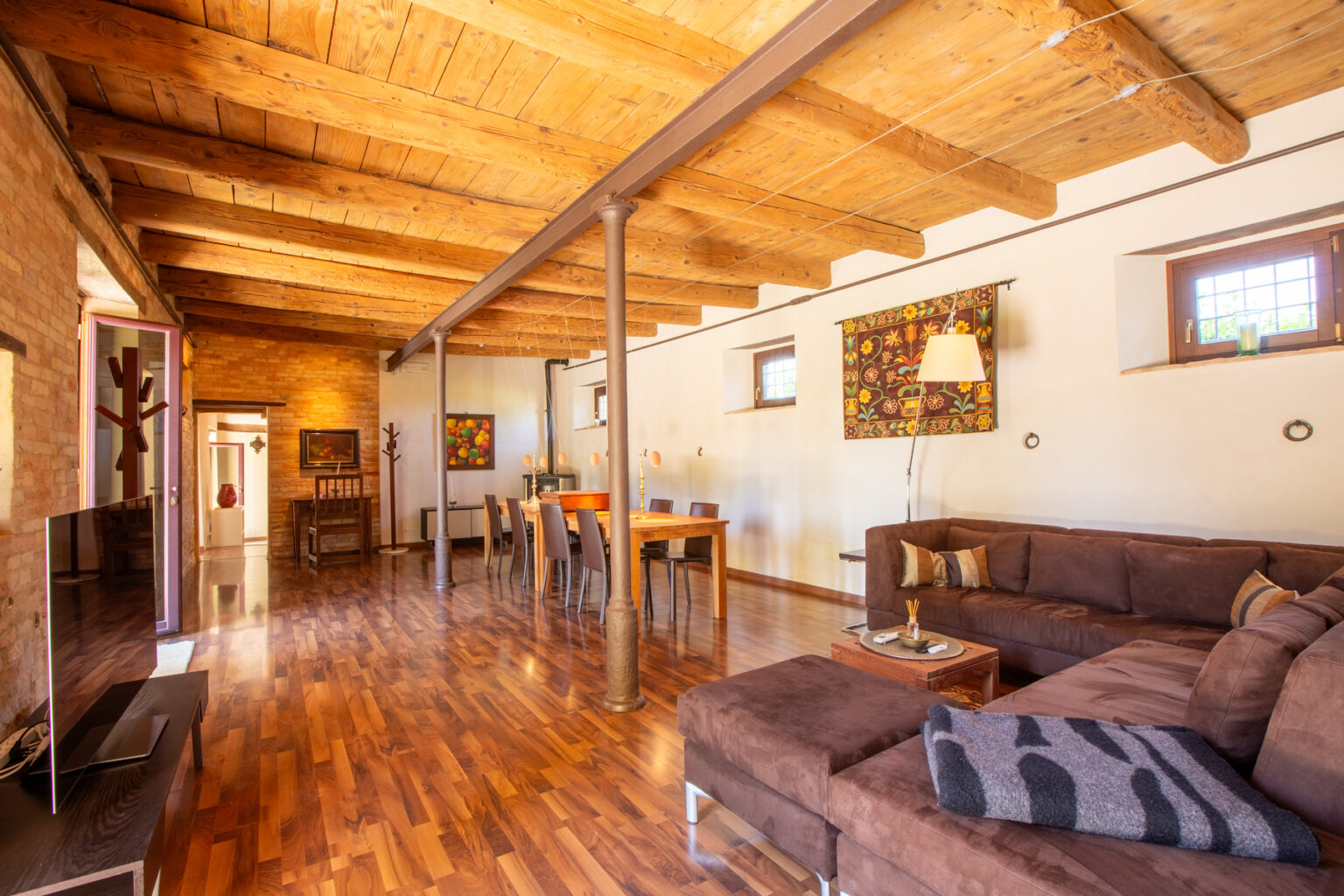 Spacious living room with wooden beams, hardwood floors, and a cozy sectional sofa, featuring a dining area with stylish decor and natural light from large windows. | Only on SopranoVillas