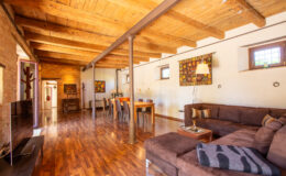 Spacious living room with wooden beams, hardwood floors, and a cozy sectional sofa, featuring a dining area with stylish decor and natural light from large windows.