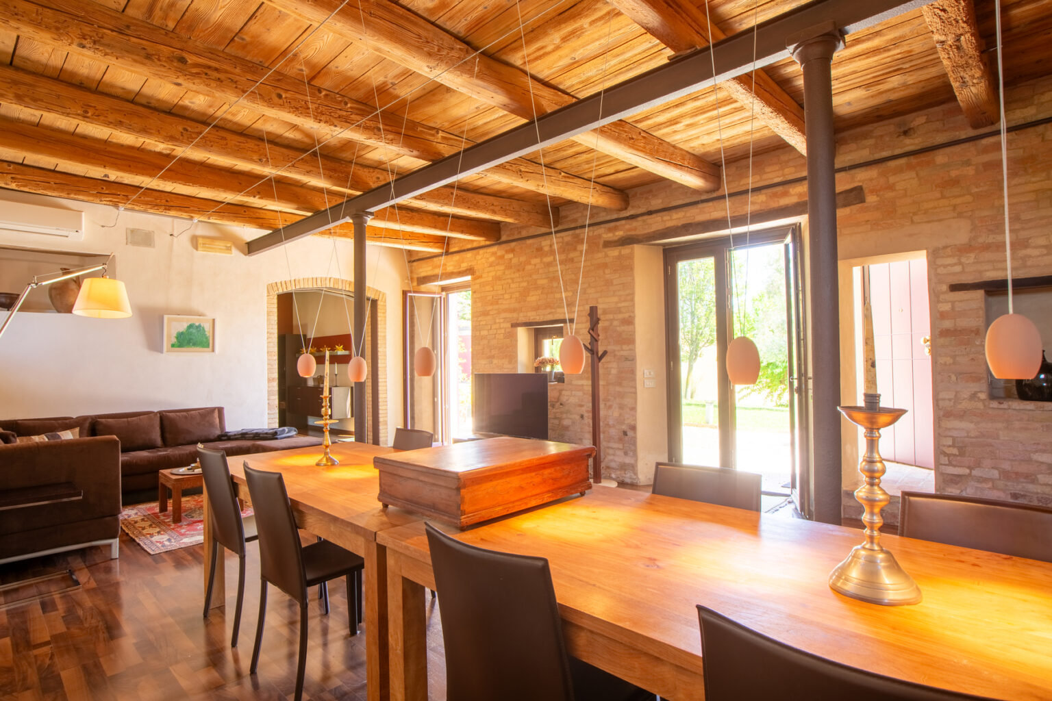 Spacious modern living room and dining area featuring exposed wooden beams, a large wooden dining table, and ample natural light from floor-to-ceiling windows. | Only on SopranoVillas