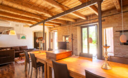 Spacious modern living room and dining area featuring exposed wooden beams, a large wooden dining table, and ample natural light from floor-to-ceiling windows.