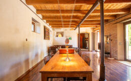 Spacious dining area featuring a wooden table, stylish chairs, and exposed wooden beams, with natural light streaming through large windows, creating a warm and inviting atmosphere.