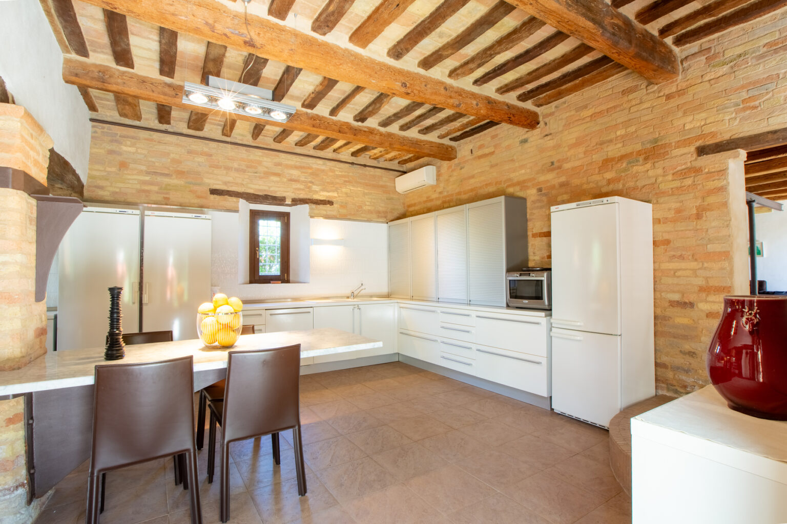 Modern kitchen with wooden beams and brick walls, featuring sleek white cabinetry, stainless steel appliances, and a dining area with a table and chairs. | Only on SopranoVillas