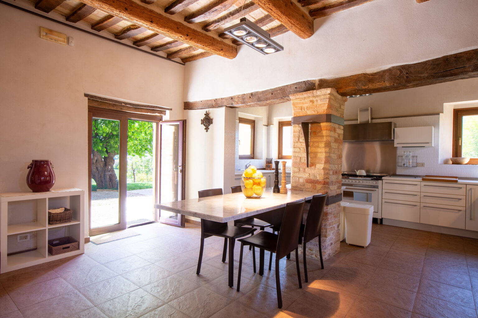 Bright and airy kitchen dining area featuring a wooden table surrounded by chairs, large windows with views of greenery, rustic wooden beams, and modern appliances. Ideal for family gatherings and meals. | Only on SopranoVillas