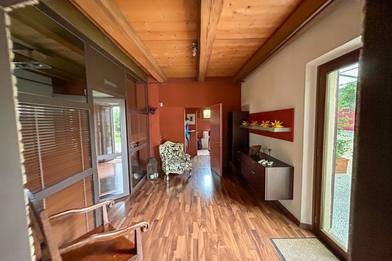 Interior view of a modern hallway featuring wooden flooring, a cozy armchair, and a decorative wooden console. Natural light streams in through large windows, highlighting the warm color palette and stylish decor. | Only on SopranoVillas