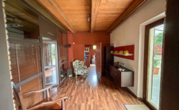 Interior view of a modern hallway featuring wooden flooring, a cozy armchair, and a decorative wooden console. Natural light streams in through large windows, highlighting the warm color palette and stylish decor.