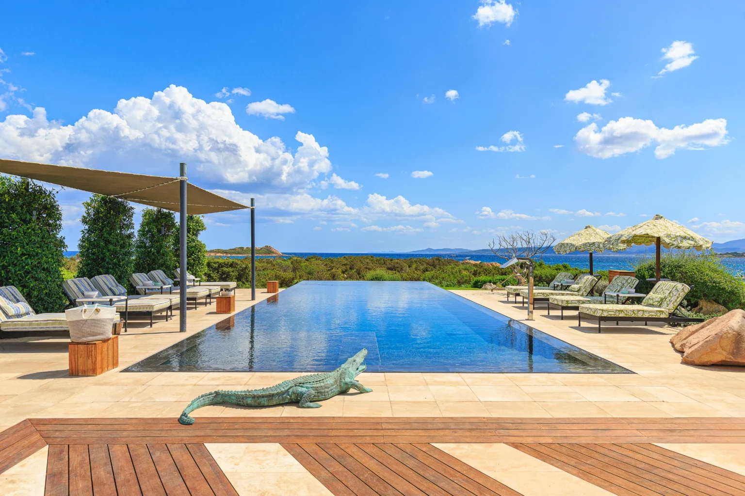 Luxury infinity pool overlooking the ocean, surrounded by lush greenery, sun loungers, and shaded umbrellas, set against a bright blue sky with fluffy clouds. A decorative crocodile sculpture adds a unique touch to the tranquil outdoor space. | Only on SopranoVillas