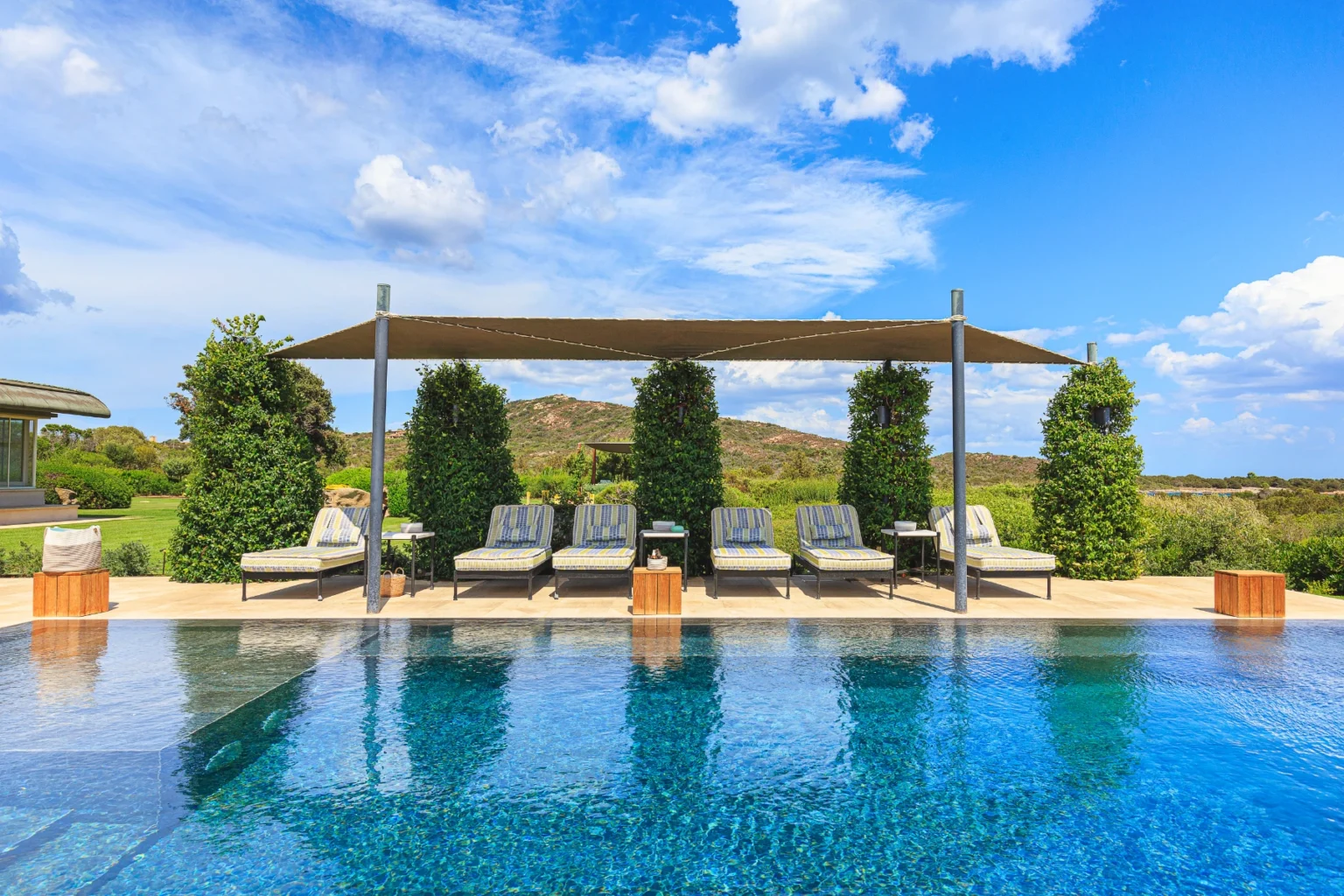 Luxury outdoor pool area featuring sun loungers under a shaded pergola, surrounded by lush greenery and scenic hills under a bright blue sky. Ideal for relaxation and leisure. | Only on SopranoVillas