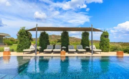 Luxury outdoor pool area featuring sun loungers under a shaded pergola, surrounded by lush greenery and scenic hills under a bright blue sky. Ideal for relaxation and leisure.