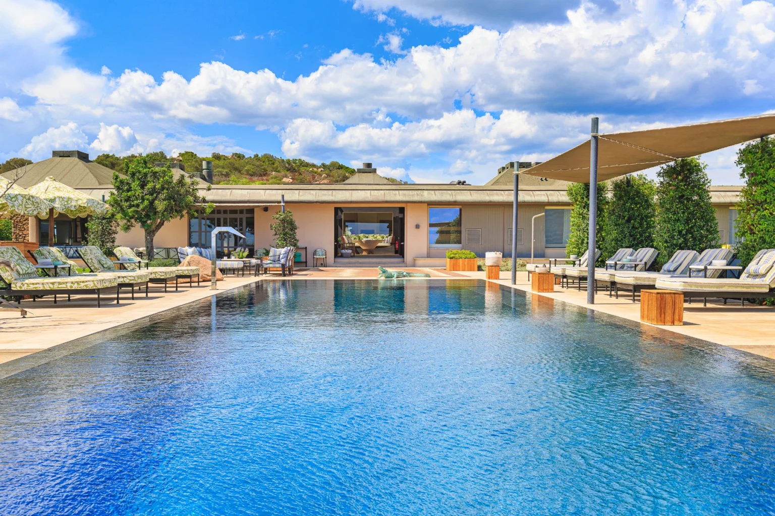 Luxurious outdoor swimming pool surrounded by comfortable lounge chairs and shaded areas, with a modern building in the background and a clear blue sky dotted with clouds. Ideal for relaxation and leisure. | Only on SopranoVillas