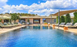 Luxurious outdoor swimming pool surrounded by comfortable lounge chairs and shaded areas, with a modern building in the background and a clear blue sky dotted with clouds. Ideal for relaxation and leisure.
