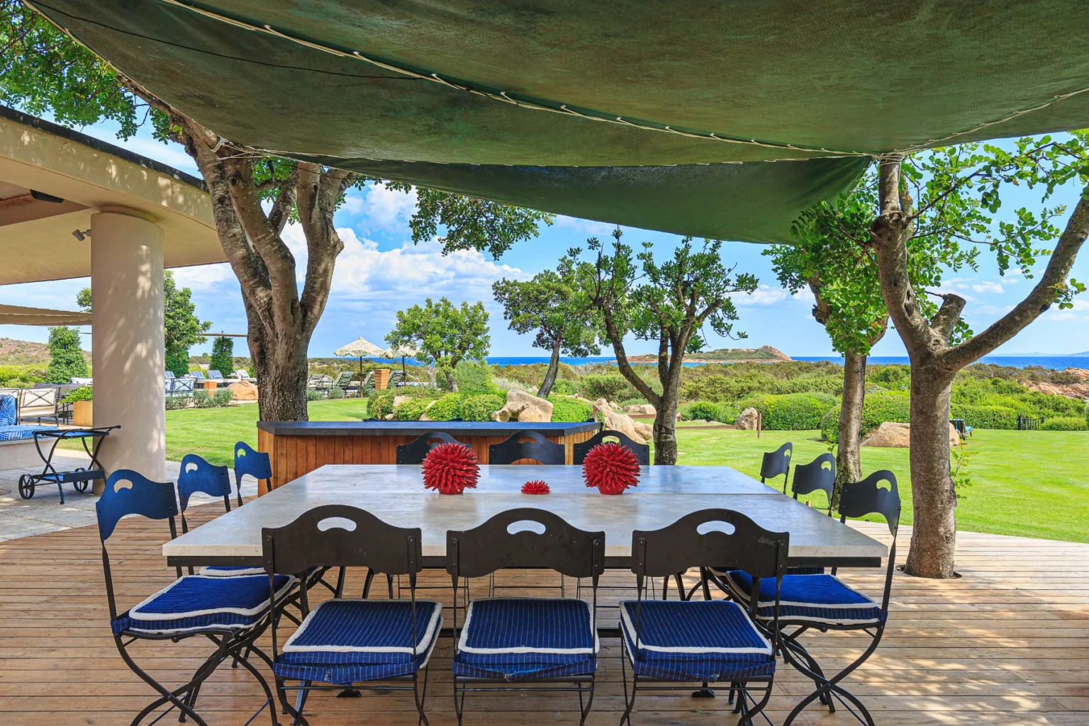 Outdoor dining area featuring a large table with blue and white striped chairs, set against a backdrop of lush greenery and ocean views. Sunlight filters through a green canopy, creating a serene atmosphere ideal for gatherings. | Only on SopranoVillas