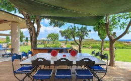 Outdoor dining area featuring a large table with blue and white striped chairs, set against a backdrop of lush greenery and ocean views. Sunlight filters through a green canopy, creating a serene atmosphere ideal for gatherings.