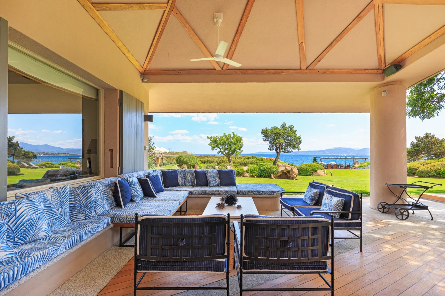 Spacious outdoor patio with comfortable seating and a view of the ocean, featuring a stylish design with blue and white cushions, wooden ceiling beams, and lush greenery in the background. Ideal for relaxation and entertaining. | Only on SopranoVillas