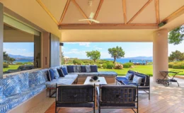 Spacious outdoor patio with comfortable seating and a view of the ocean, featuring a stylish design with blue and white cushions, wooden ceiling beams, and lush greenery in the background. Ideal for relaxation and entertaining.