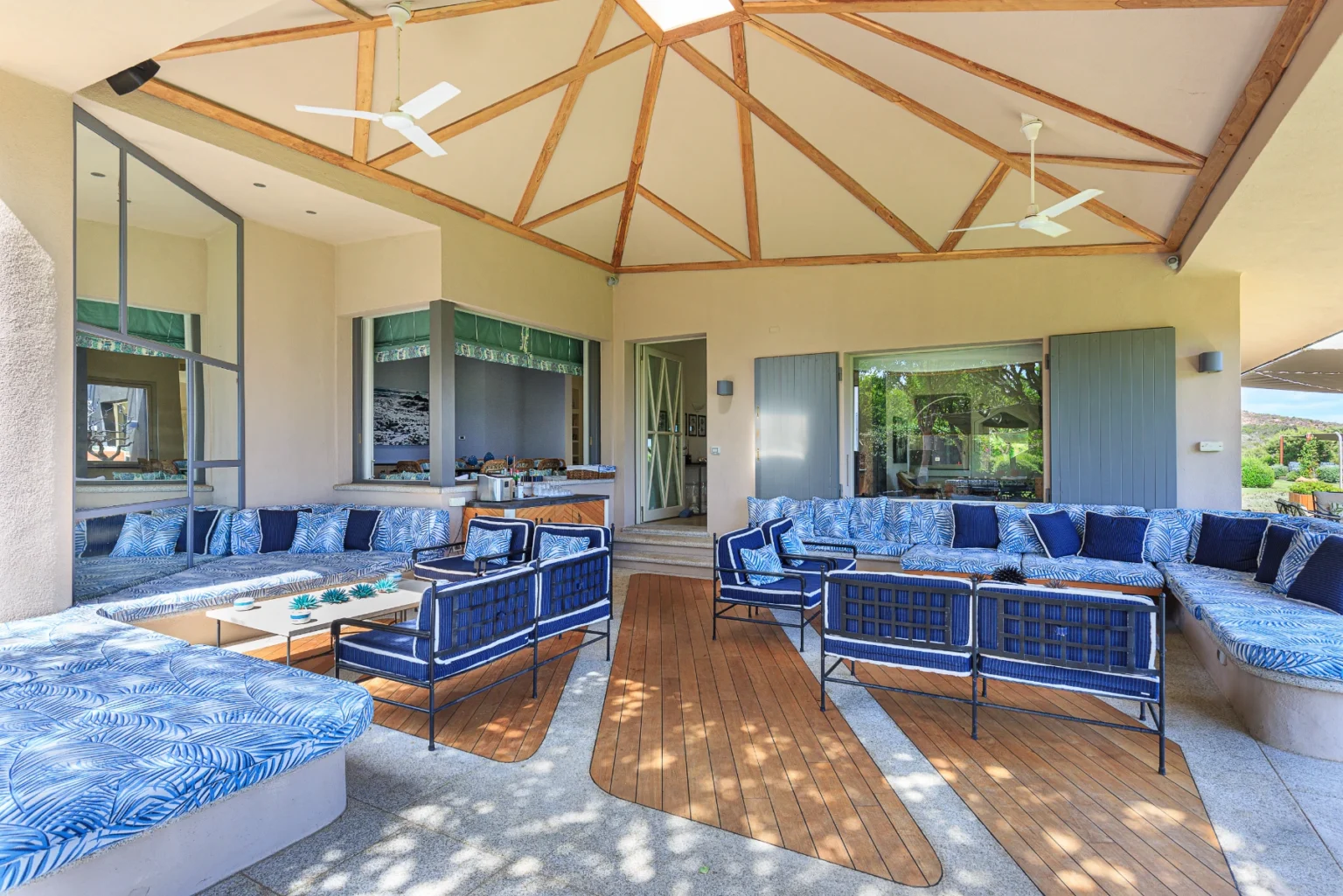 Spacious outdoor living area featuring a stylish seating arrangement with blue patterned cushions, a wooden deck, and a ceiling fan. The design includes large windows and a bright, airy atmosphere, ideal for relaxation and entertaining. | Only on SopranoVillas
