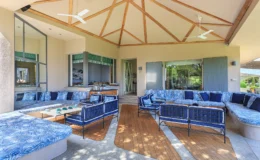 Spacious outdoor living area featuring a stylish seating arrangement with blue patterned cushions, a wooden deck, and a ceiling fan. The design includes large windows and a bright, airy atmosphere, ideal for relaxation and entertaining.