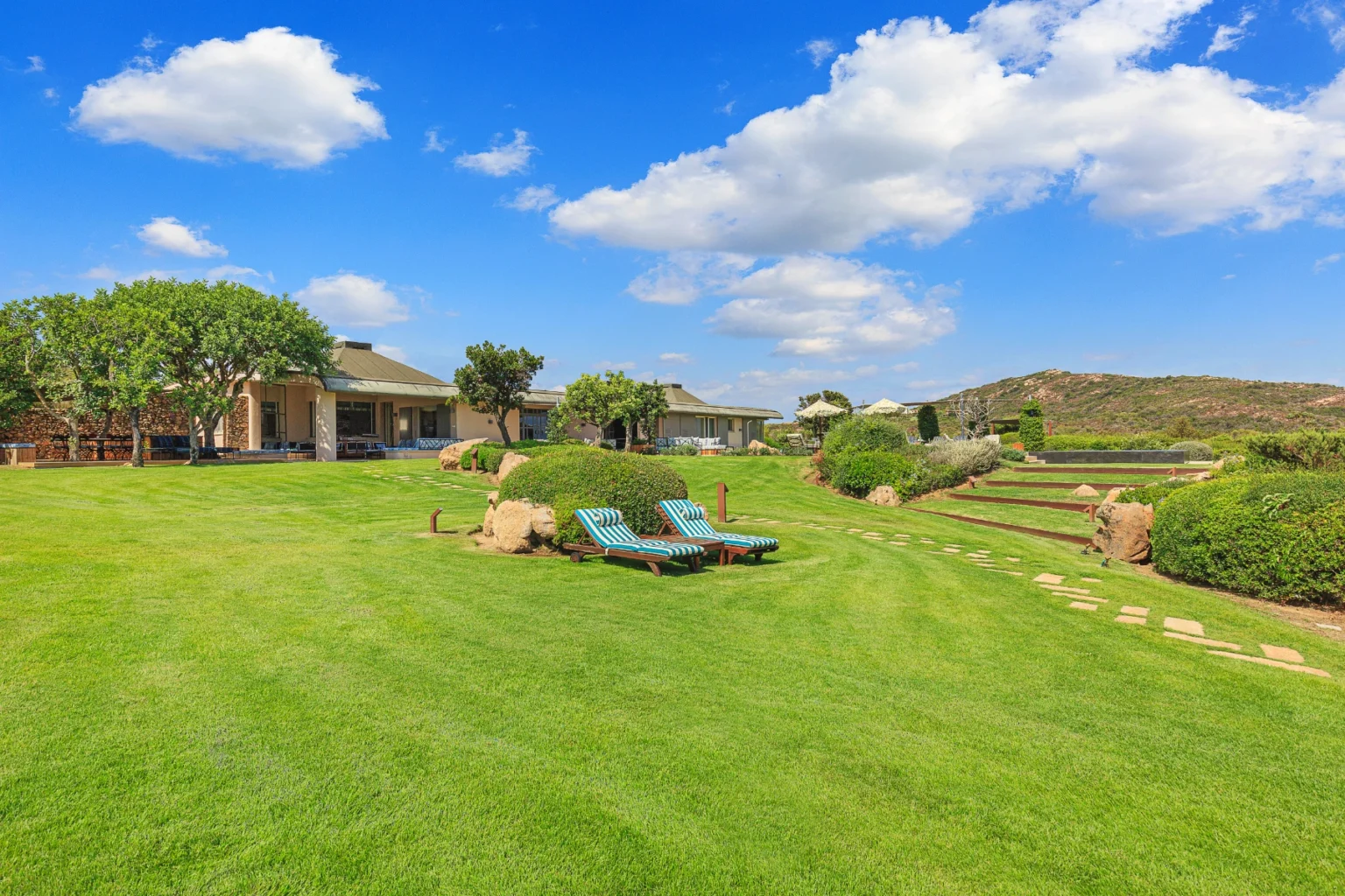Spacious backyard of a modern home featuring well-manicured green lawn, comfortable lounge chairs, and landscaped gardens under a bright blue sky with fluffy clouds. | Only on SopranoVillas