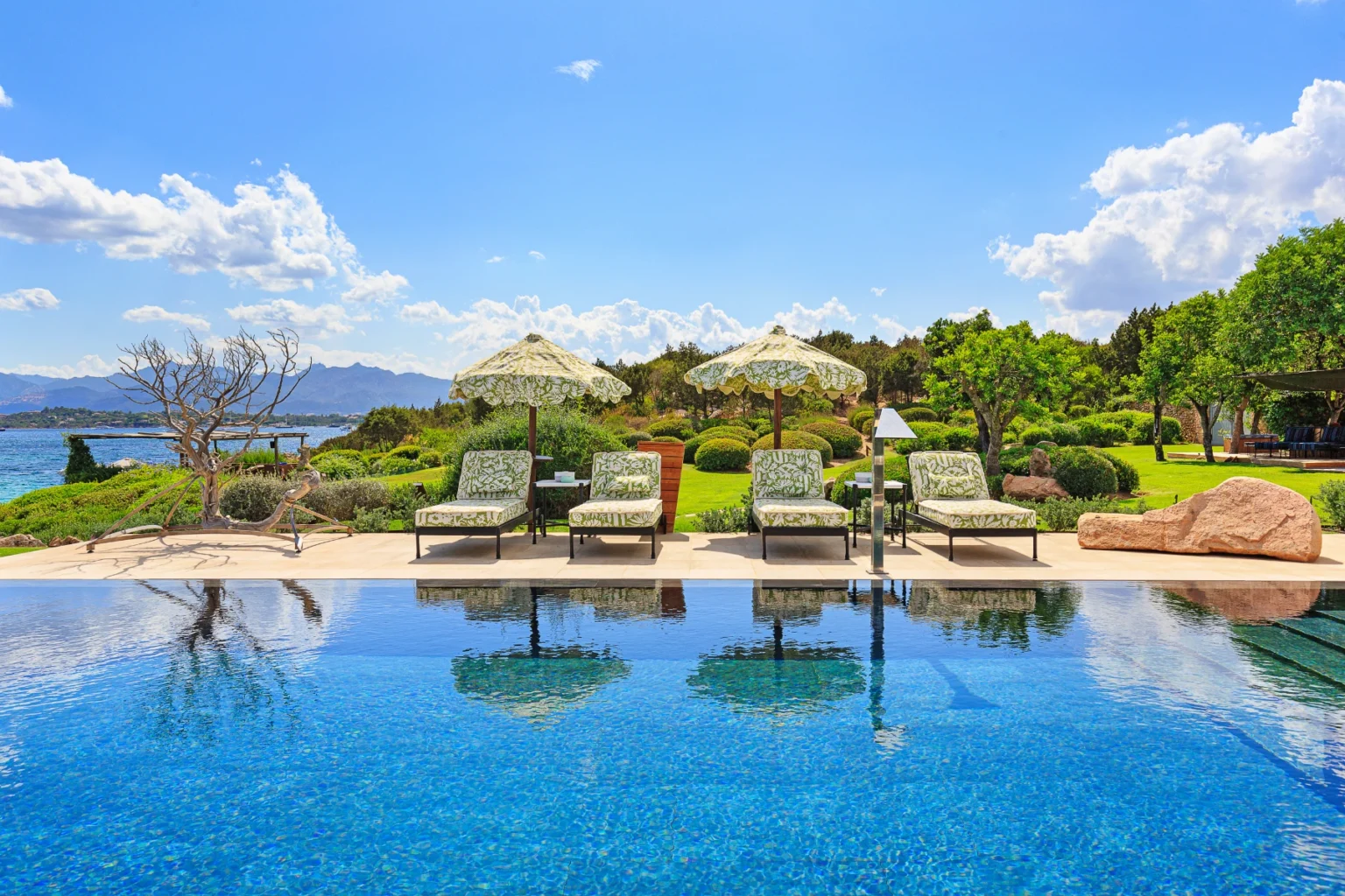 Luxurious outdoor pool area featuring comfortable lounge chairs with patterned cushions, shaded by decorative umbrellas, overlooking a scenic landscape of lush greenery and mountains under a bright blue sky. | Only on SopranoVillas
