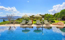 Luxurious outdoor pool area featuring comfortable lounge chairs with patterned cushions, shaded by decorative umbrellas, overlooking a scenic landscape of lush greenery and mountains under a bright blue sky.