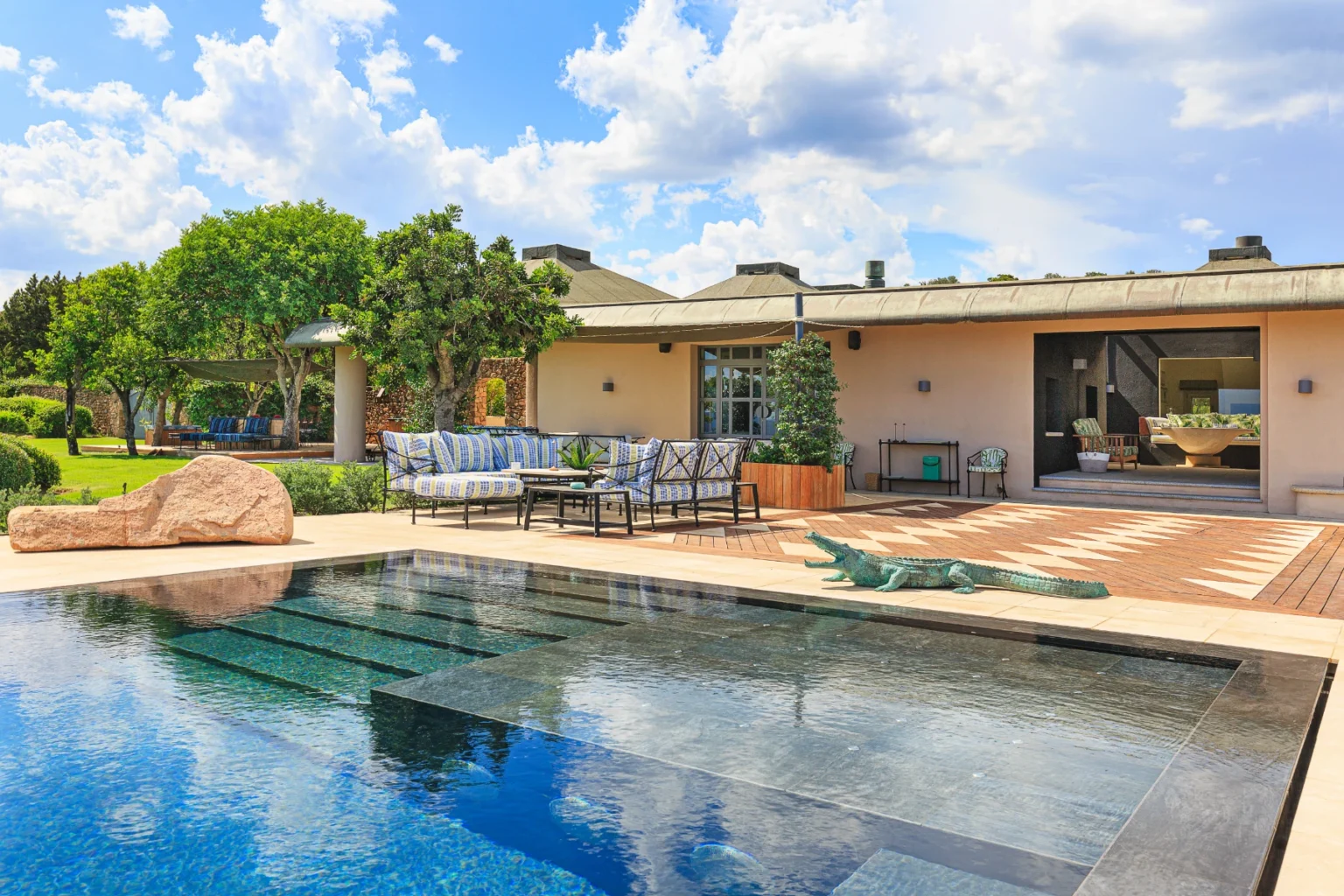 Luxurious outdoor living space featuring a modern swimming pool, stylish seating area with blue and white striped cushions, and landscaped gardens under a bright blue sky with fluffy clouds. The elegant architecture and natural elements create a serene and inviting atmosphere. | Only on SopranoVillas
