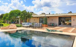 Luxurious outdoor living space featuring a modern swimming pool, stylish seating area with blue and white striped cushions, and landscaped gardens under a bright blue sky with fluffy clouds. The elegant architecture and natural elements create a serene and inviting atmosphere.