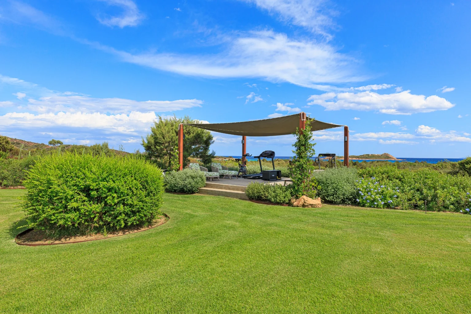 Lush green lawn with a shaded outdoor seating area overlooking the ocean, featuring comfortable loungers and fitness equipment, under a bright blue sky with scattered clouds. | Only on SopranoVillas