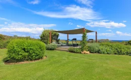 Lush green lawn with a shaded outdoor seating area overlooking the ocean, featuring comfortable loungers and fitness equipment, under a bright blue sky with scattered clouds.