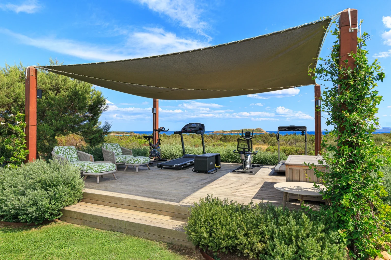 Outdoor fitness area featuring exercise equipment, including a treadmill and stationary bike, under a shaded canopy. Comfortable seating with green patterned cushions is arranged on a wooden deck, surrounded by lush greenery and a scenic view of the ocean. Ideal for outdoor workouts and relaxation. | Only on SopranoVillas