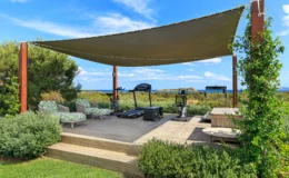 Outdoor fitness area featuring exercise equipment, including a treadmill and stationary bike, under a shaded canopy. Comfortable seating with green patterned cushions is arranged on a wooden deck, surrounded by lush greenery and a scenic view of the ocean. Ideal for outdoor workouts and relaxation.