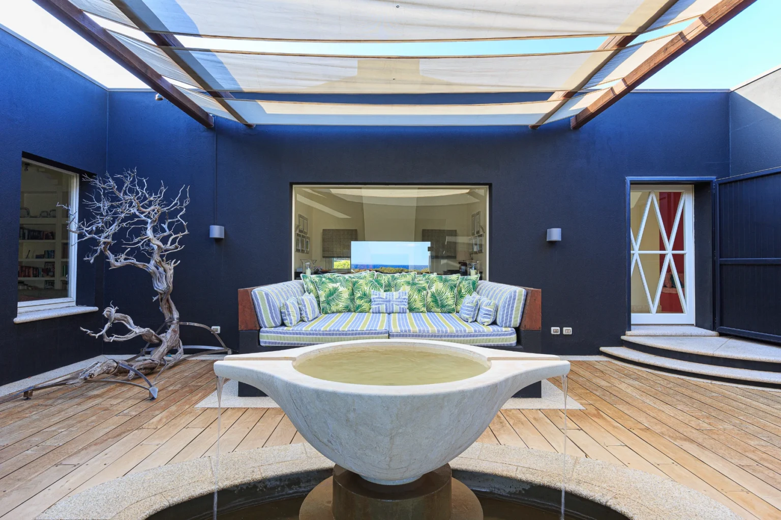 Modern outdoor patio featuring a stylish sofa with green and striped cushions, a decorative stone fountain, and a unique driftwood sculpture. The space is illuminated by natural light through large windows, with a sleek design and wooden decking, ideal for relaxation and entertaining. | Only on SopranoVillas