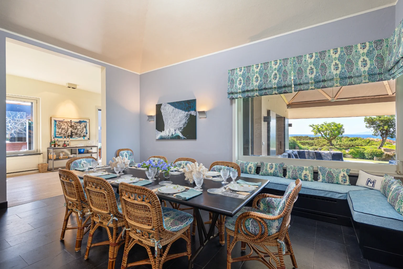 Elegant dining room featuring a large black table set for a meal, surrounded by stylish wicker chairs. The walls are painted in soft blue, complemented by patterned window treatments. A vibrant floral centerpiece adds color, while a large window offers scenic views of the outdoors. A modern artwork hangs on the wall, enhancing the room's contemporary decor. | Only on SopranoVillas