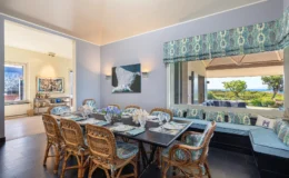 Elegant dining room featuring a large black table set for a meal, surrounded by stylish wicker chairs. The walls are painted in soft blue, complemented by patterned window treatments. A vibrant floral centerpiece adds color, while a large window offers scenic views of the outdoors. A modern artwork hangs on the wall, enhancing the room's contemporary decor.