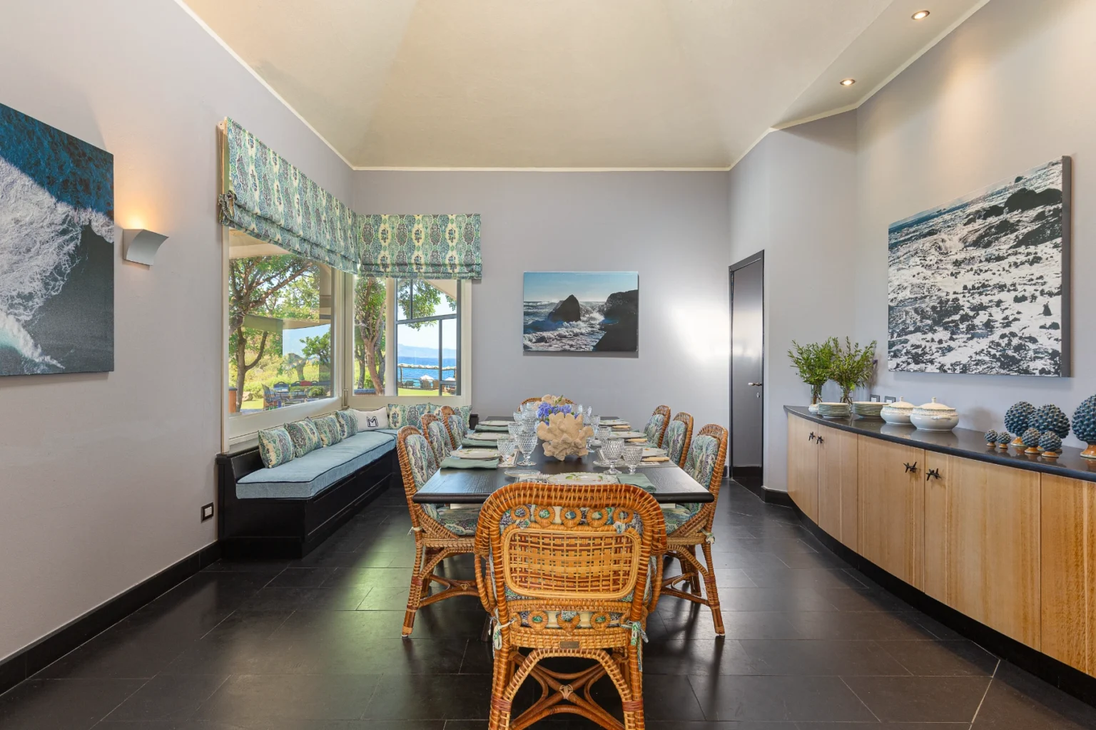 Spacious dining room featuring a long wooden table set for a meal, surrounded by rattan chairs with decorative cushions. Large windows provide a view of the outdoors, and coastal-themed artwork adorns the walls. A cozy window seat and a sideboard with decorative items enhance the room's inviting atmosphere. | Only on SopranoVillas