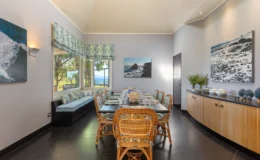 Spacious dining room featuring a long wooden table set for a meal, surrounded by rattan chairs with decorative cushions. Large windows provide a view of the outdoors, and coastal-themed artwork adorns the walls. A cozy window seat and a sideboard with decorative items enhance the room's inviting atmosphere.