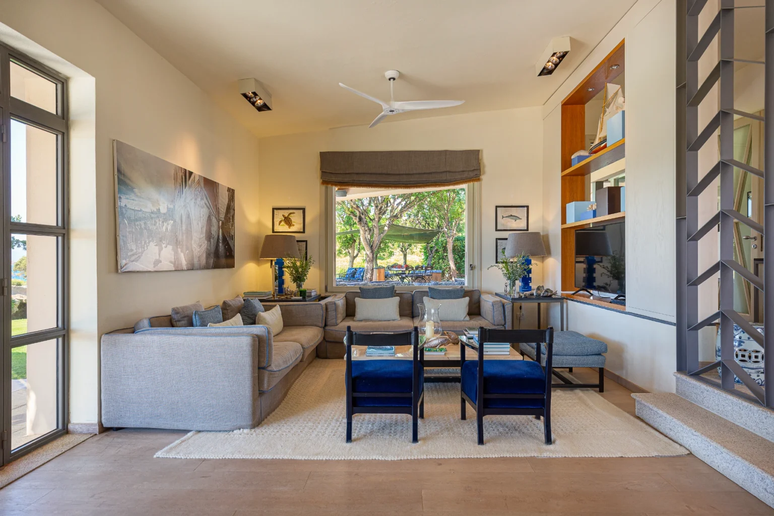 Spacious and modern living room featuring a comfortable gray sectional sofa, blue accent chairs, and a large window with a view of the outdoors. Decorated with artwork, a coffee table, and stylish lamps, the room exudes a relaxed ambiance perfect for entertaining or unwinding. | Only on SopranoVillas