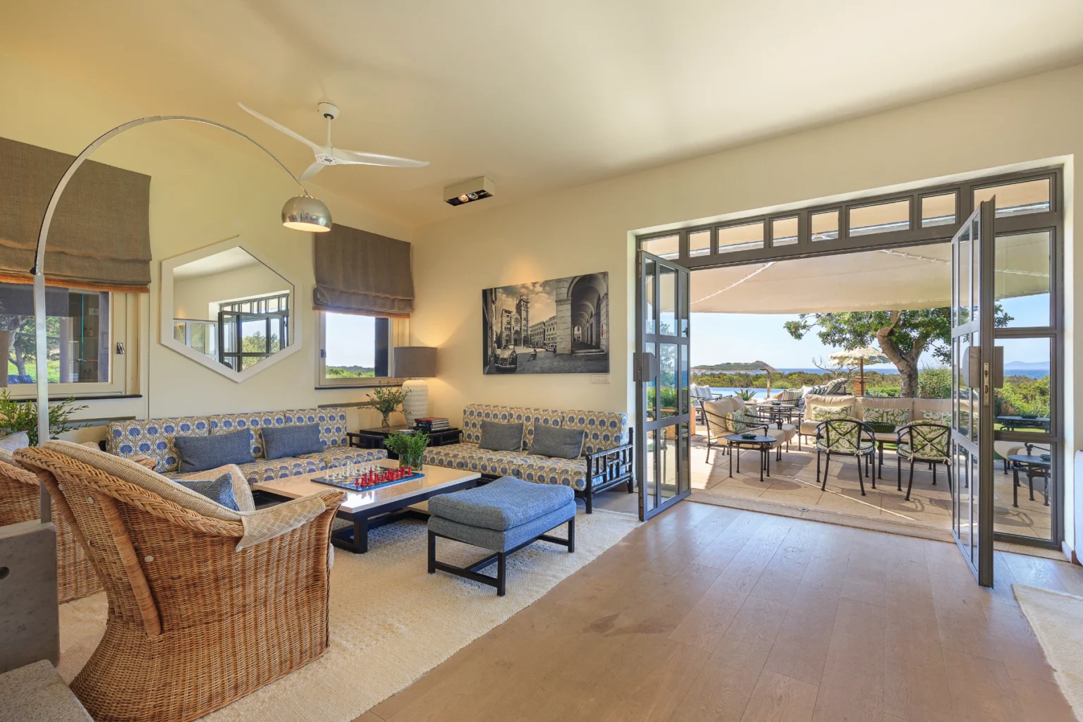 Spacious and bright living room featuring comfortable seating, a modern ceiling fan, and large windows that open to a scenic outdoor patio. The decor includes a stylish coffee table with a chess set and artwork on the walls, creating a welcoming atmosphere for relaxation and entertainment. | Only on SopranoVillas