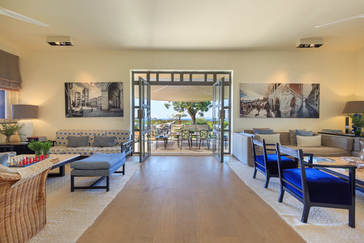 Spacious modern living room with stylish furniture, featuring a patterned sofa, comfortable chairs, and large windows that open to a scenic outdoor terrace. Decorated with artwork and a chess set, the room offers a bright and inviting atmosphere. | Only on SopranoVillas