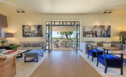 Spacious modern living room with stylish furniture, featuring a patterned sofa, comfortable chairs, and large windows that open to a scenic outdoor terrace. Decorated with artwork and a chess set, the room offers a bright and inviting atmosphere.