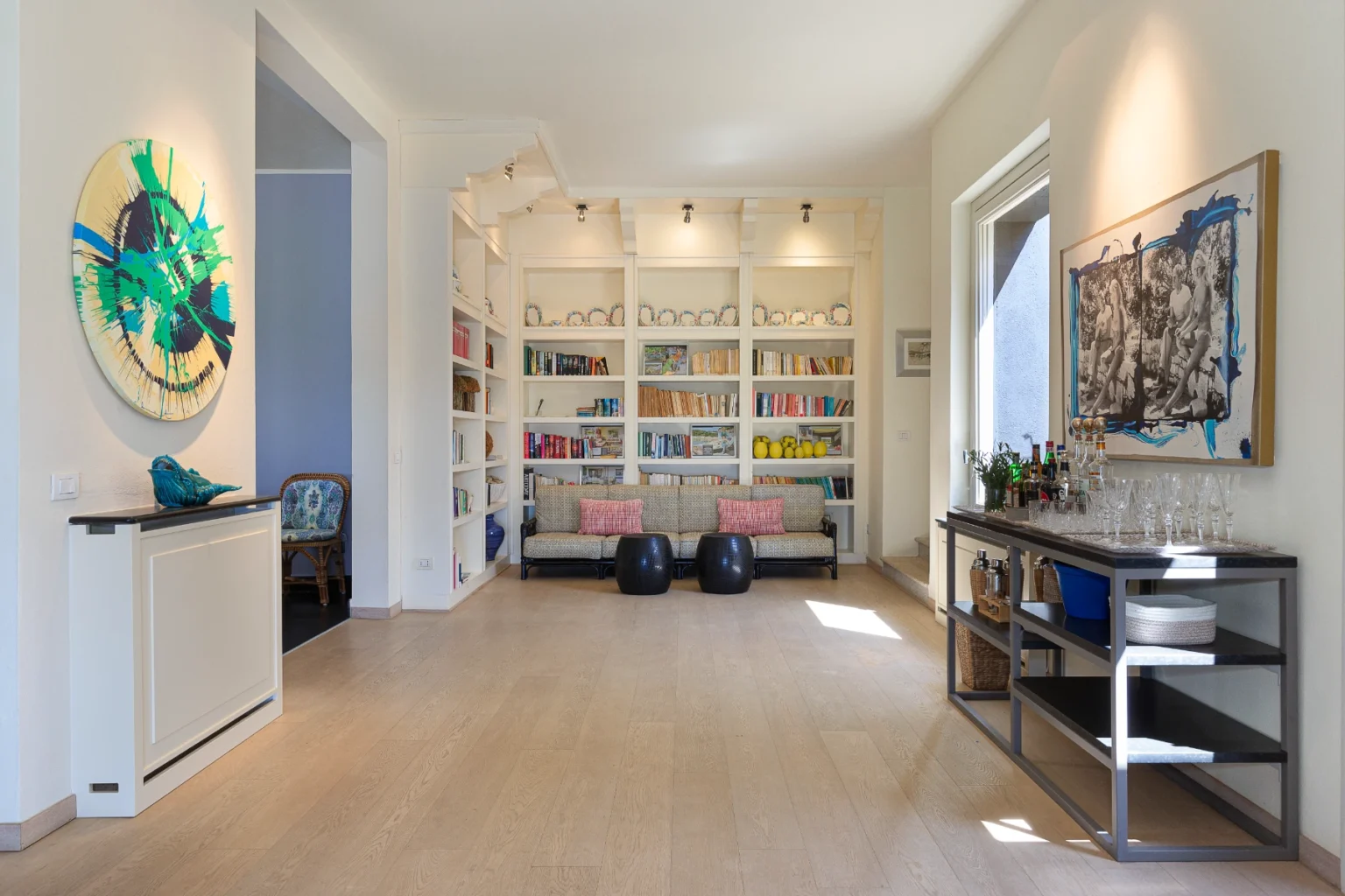 Spacious modern living room featuring a colorful abstract painting, a stylish bookshelf filled with books, a comfortable sofa with decorative pillows, and a sleek bar cart with glassware and beverages. Natural light floods the space through large windows, enhancing the warm wood flooring and inviting atmosphere. | Only on SopranoVillas
