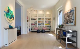 Spacious modern living room featuring a colorful abstract painting, a stylish bookshelf filled with books, a comfortable sofa with decorative pillows, and a sleek bar cart with glassware and beverages. Natural light floods the space through large windows, enhancing the warm wood flooring and inviting atmosphere.