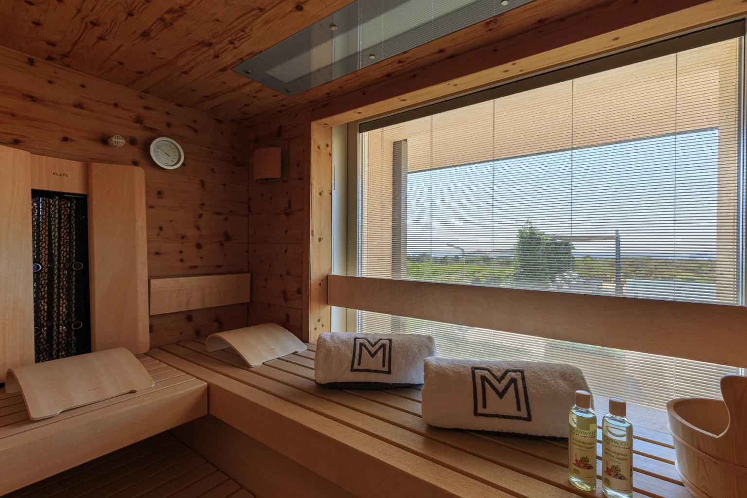 Interior of a modern sauna featuring wooden benches, towels with a logo, a clock, and a view through large windows with blinds. The space is designed for relaxation, with essential oils placed on a bench for aromatherapy. | Only on SopranoVillas